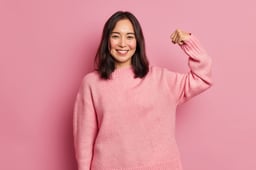 powerful-cheerful-brunette-woman-raises-arm-shows-muscle-demonstrates-her-strength-looks-confident-camera-smiles-gently-wears-casual-long-sleeved-jumper